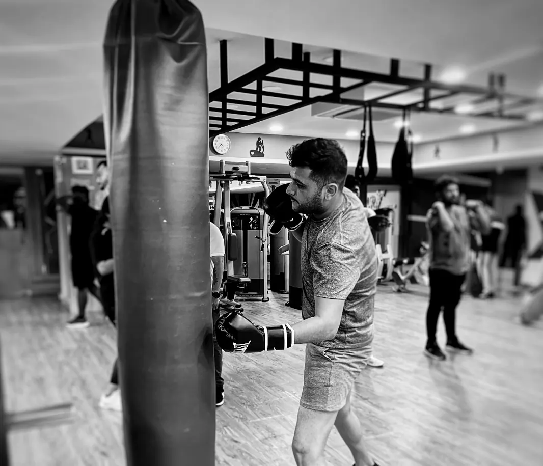 Black and white photo of a man in boxing gloves punching a heavy bag, other members training behind him