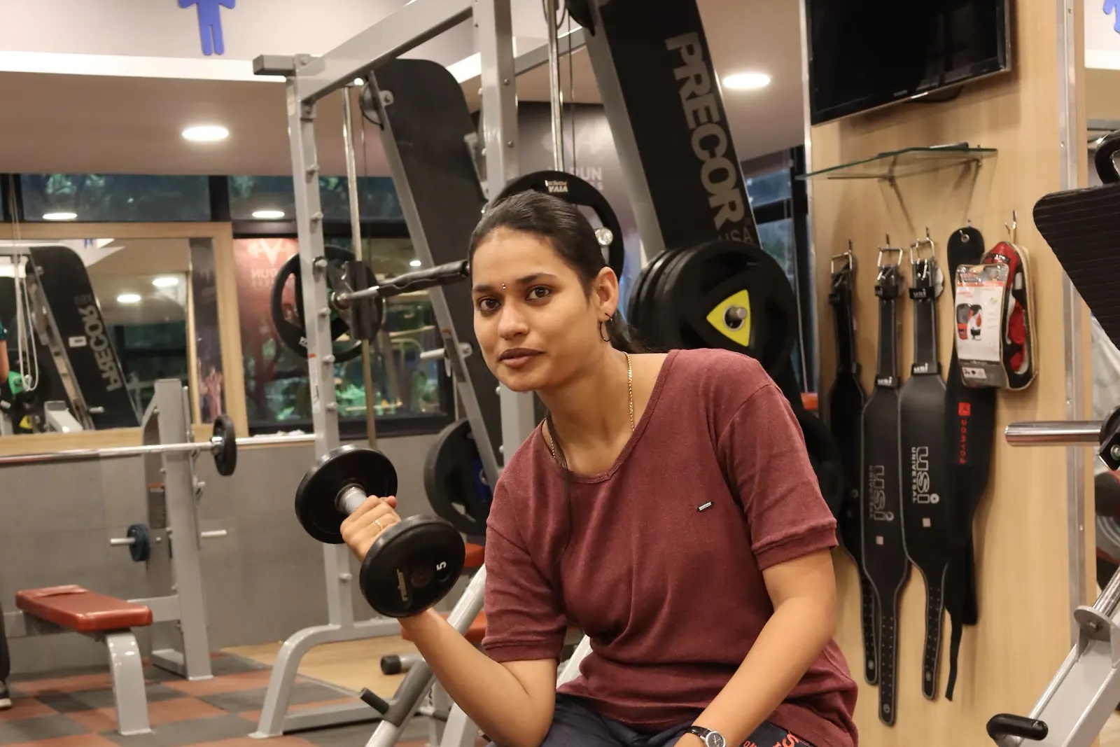 A woman in a maroon t-shirt sits on a bench curling a dumbbell