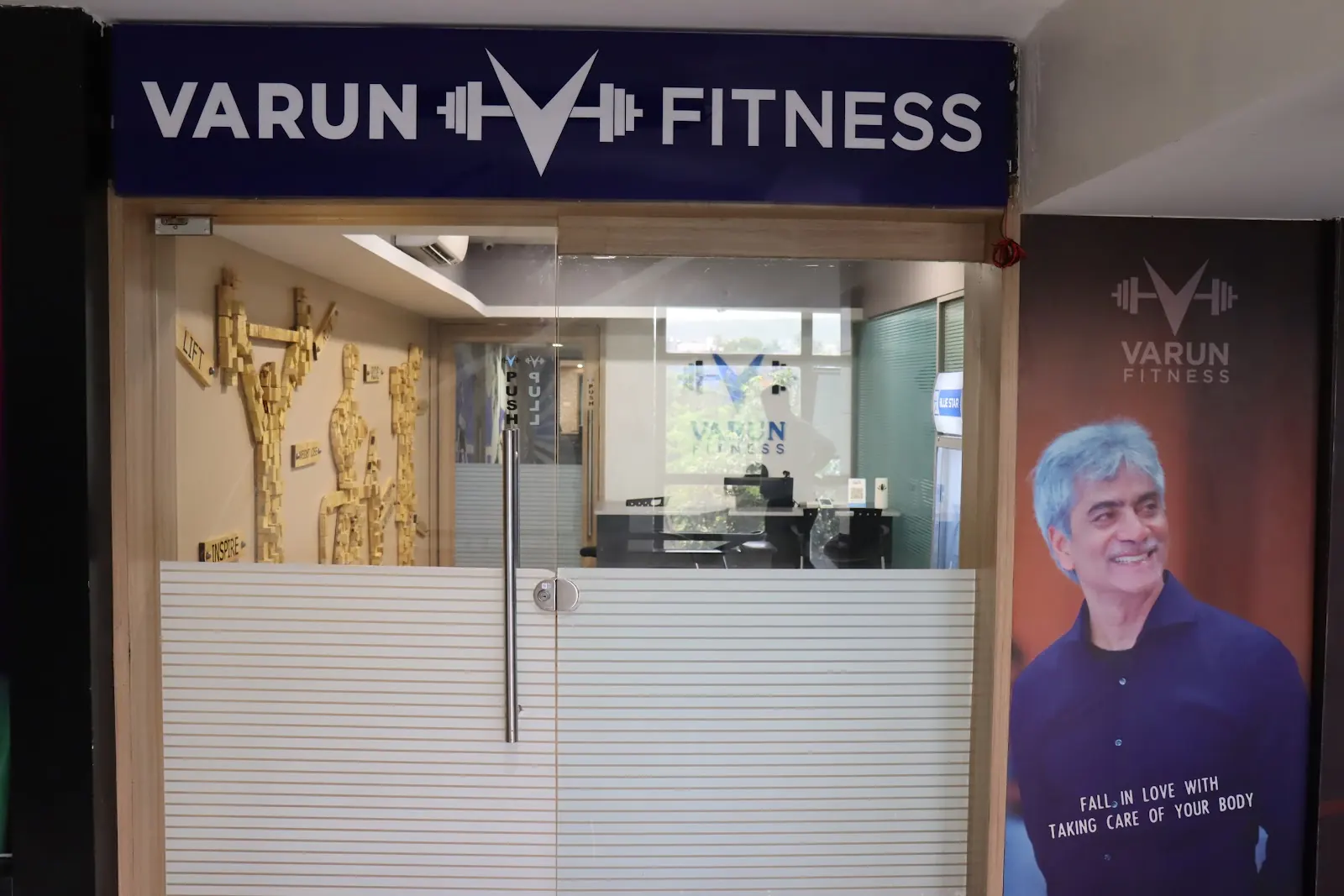 Glass entrance with a Varun Fitness signboard above it and a poster beside the door