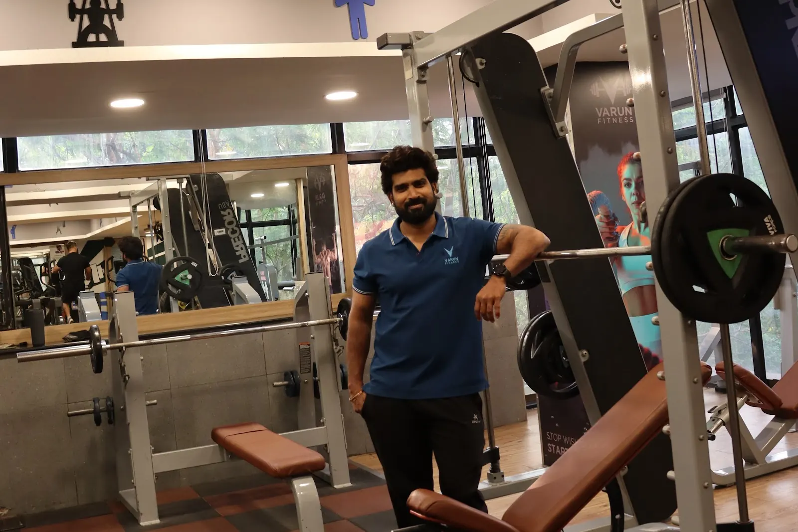 A man in a blue polo stands with his arm on a loaded barbell in a smith machine