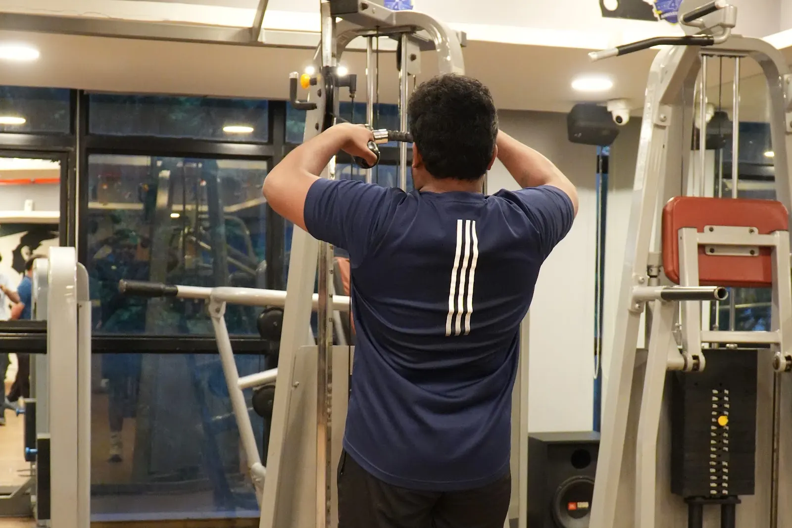 A man in a navy t-shirt, seen from behind, pulls the bar on a cable machine