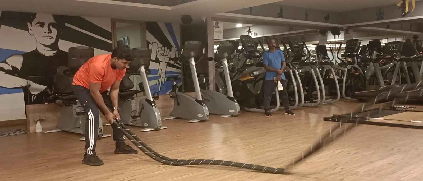 A man in an orange top pulls heavy battle ropes on a wooden floor, a second man and rows of cardio machines behind him
